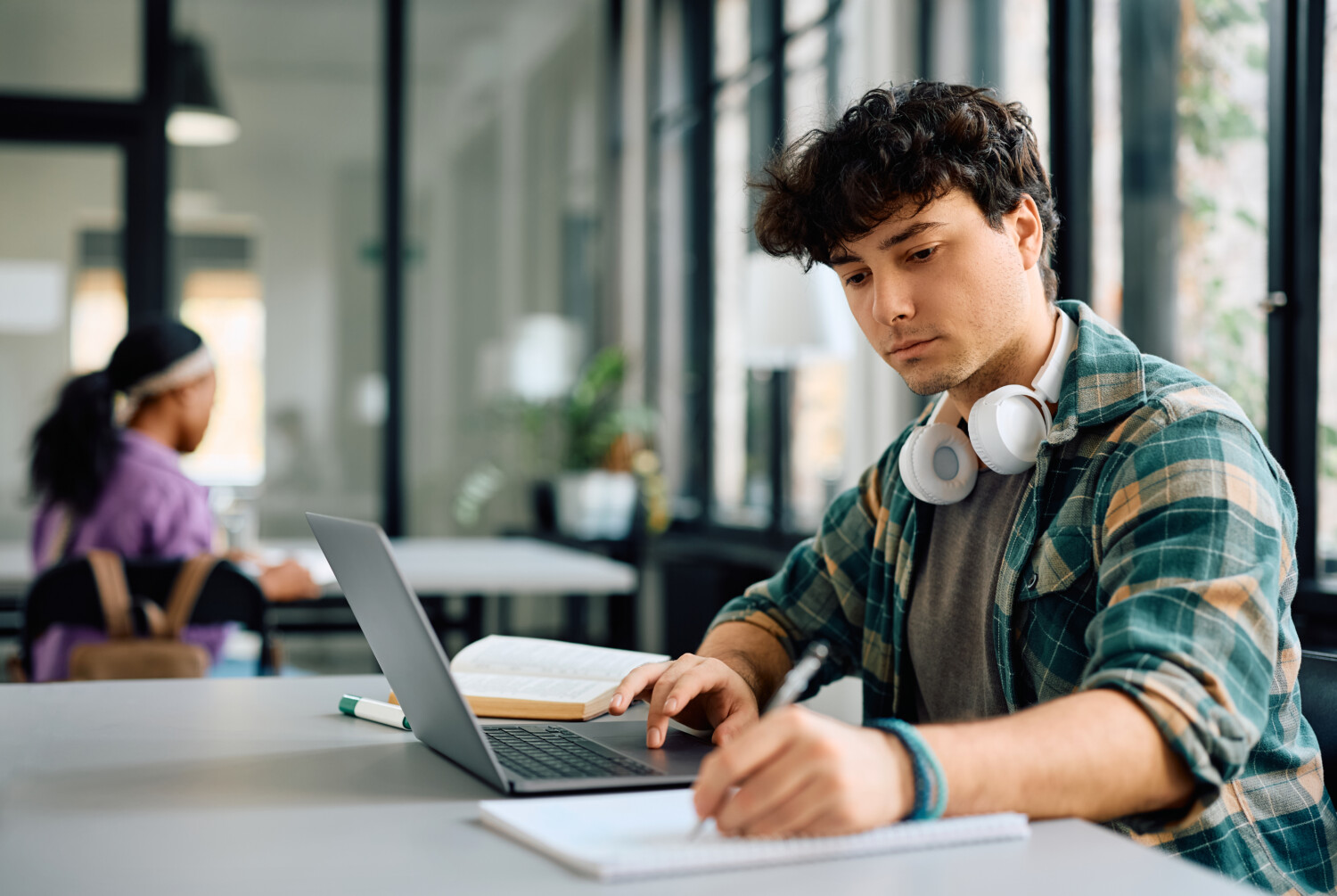 A student at a desk with a laptop, taking notes in a notepad