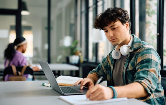 A student at a desk with a laptop, taking notes in a notepad